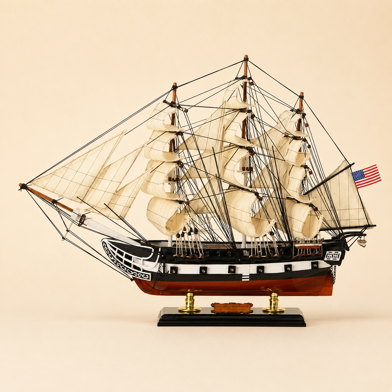 Side view of an American three-masted tall ship model featuring layered cream sails, intricate black rope rigging, black and white hull, red lower hull and polished black presentation base.