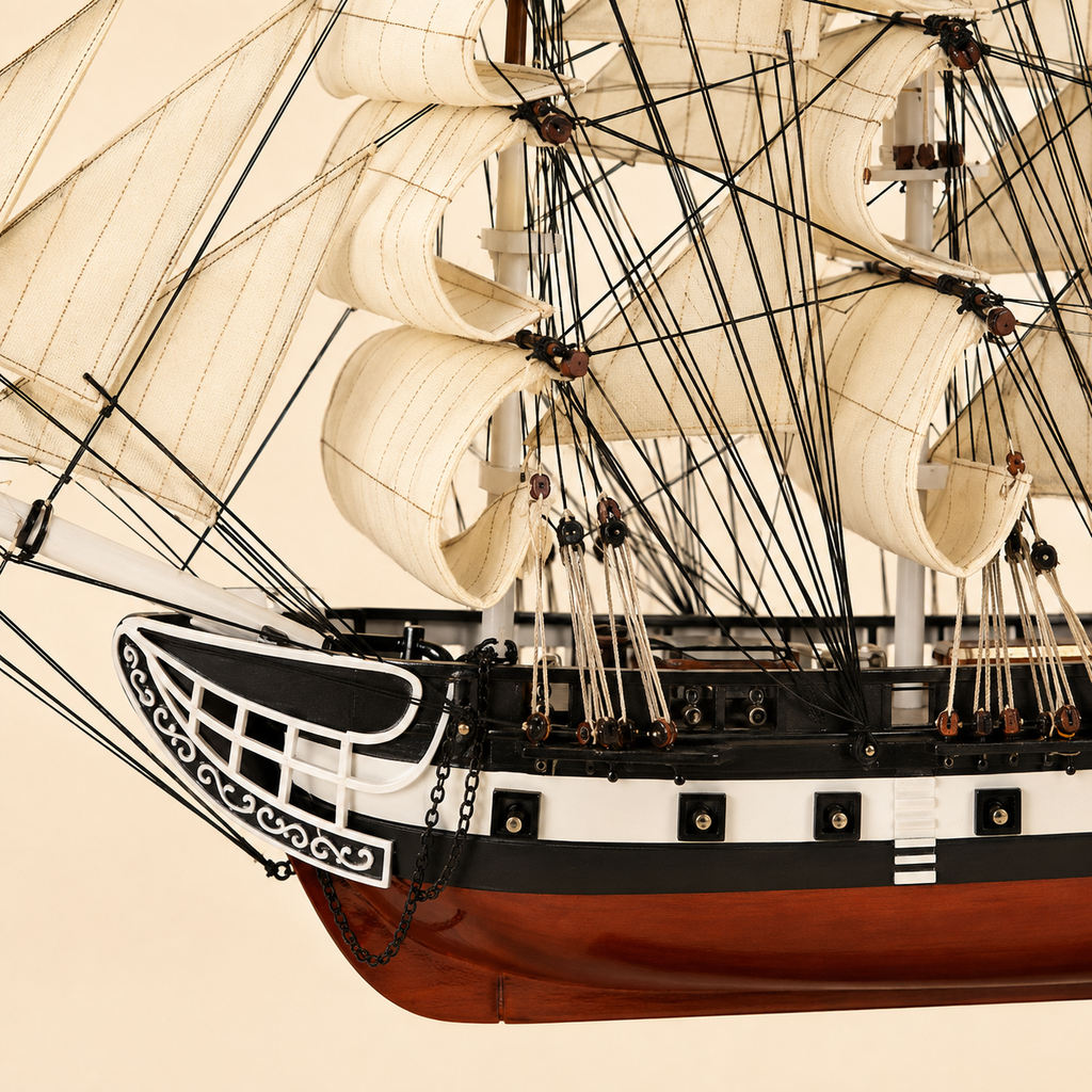 Close-up of the American tall ship model showing textured cream sails, black rigging, wooden pulleys, rope knots, deck railings, ornate bow details, anchor chains and handcrafted hull finishes.