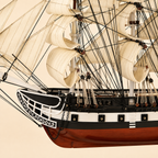 Close-up of the American tall ship model showing textured cream sails, black rigging, wooden pulleys, rope knots, deck railings, ornate bow details, anchor chains and handcrafted hull finishes.