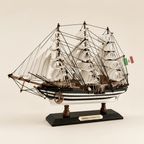 Vista lateral del modelo de gran velero Amerigo Vespucci de la Marina italiana sobre fondo crema, con silueta completa de tres mástiles, velas blancas completas, densa jarcia negra, casco negro y blanco a rayas con puntos blancos, bandera de la Marina italiana, ancla en proa y base negra con placa dorada