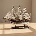 Modelo de gran velero Amerigo Vespucci de la Marina italiana sobre un escritorio de madera en un ambiente cálido del hogar, con velas blancas de aparejo completo en tres mástiles, casco negro y blanco a rayas con puntos blancos, densa jarcia negra, bandera de la Marina italiana y base negra con placa dorada AMERIGO VESPUCCI