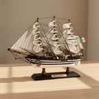 Modelo de gran velero Amerigo Vespucci de la Marina italiana sobre un escritorio de madera en un ambiente cálido del hogar, con velas blancas de aparejo completo en tres mástiles, casco negro y blanco a rayas con puntos blancos, densa jarcia negra, bandera de la Marina italiana y base negra con placa dorada AMERIGO VESPUCCI