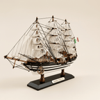 Vista frontal de tres cuartos del modelo de gran velero Amerigo Vespucci de la Marina italiana sobre fondo crema, con tres mástiles y velas blancas completas de aparejo cuadrado, densa jarcia negra, casco negro y blanco a rayas con puntos blancos, ancla en proa y base negra con placa dorada