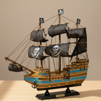  Handcrafted wooden pirate ship model displayed on a light wooden desk in a warm home setting, showing three masts with black skull-and-crossbones sails, blue-and-wood-grain striped hull with cannon ports, rope rigging, anchor at bow, and black display base with Pirate Ship nameplate – 38 cm collectible ship model