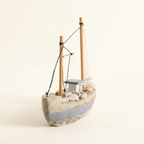 Mediterranean fishing boat wooden model front 3/4 view – wide low hull with weathered white-grey finish and grey waterline stripe, two wooden masts, black rope rigging, linen sail, rope coils and seashell deck details, cream white background